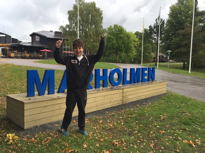 En ungdom sträcker upp armarna i luften framför en skylt med texten Marholmen.